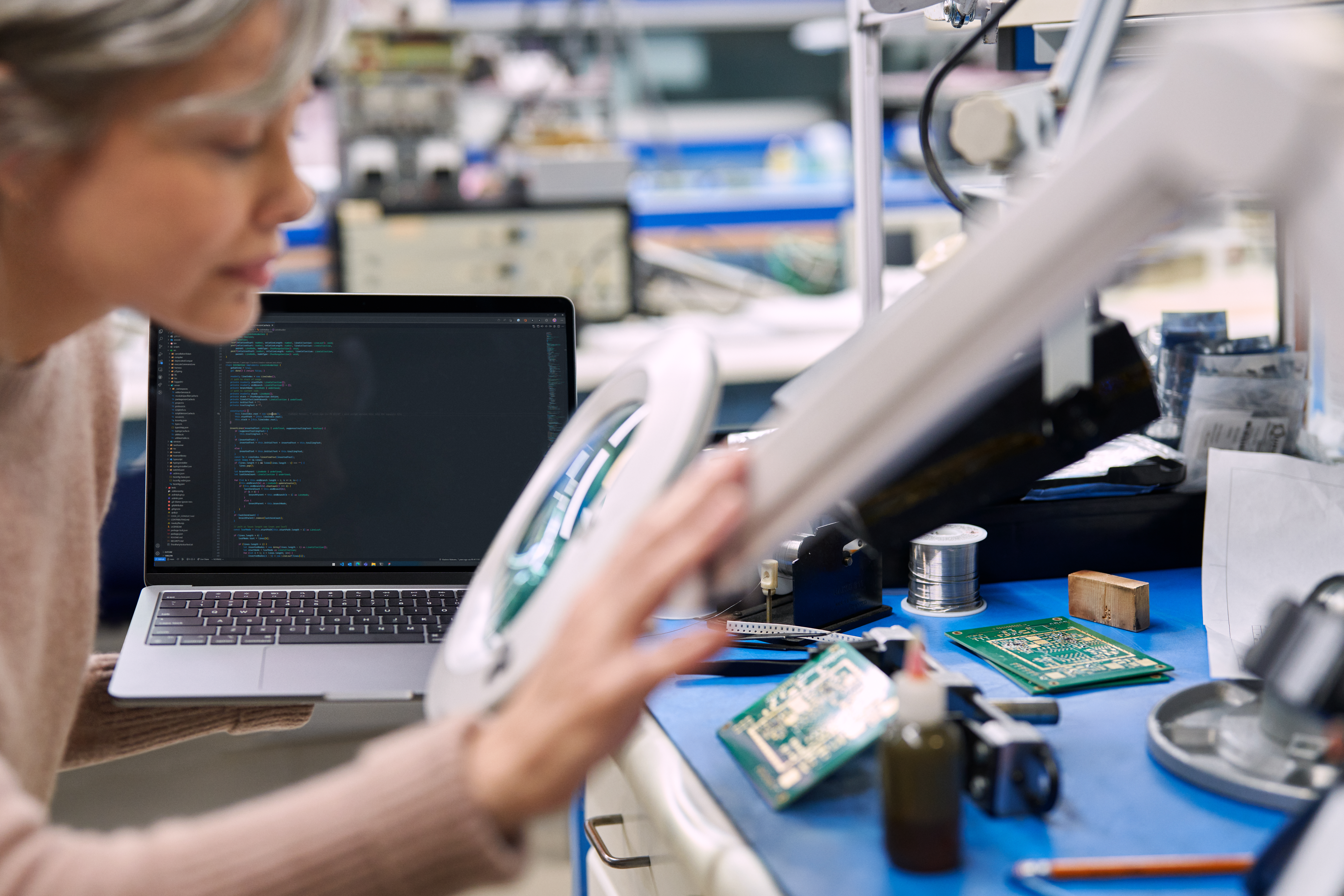 Person working at a desk with circuit boards, a magnifying lamp, and a laptop displaying code in an electronics lab.